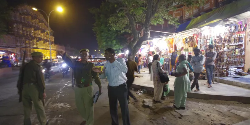 Polizei in Indien zeigt in eine Richtung. Daneben steht eine Person. Im Hintergrund sind Straßengeschäfte.