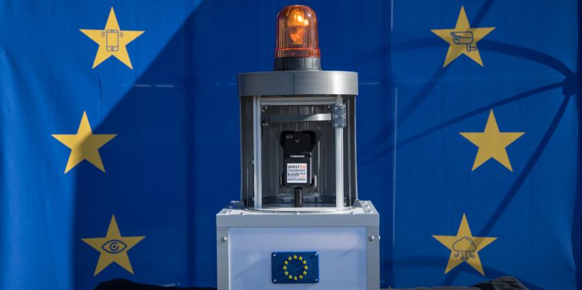Vor einer EU-Flagge mit Sternen, die Überwachungssymbole in sich haben, steht ein Apparat. Der Apparat hat mittig eine Handyhalterung, oben eine Alarmleuchte und unten ein Gehäuse aus Plexiglas mit Metallprofilen.