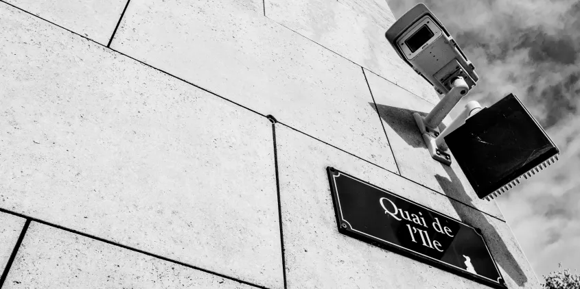 Detailaufnahme einer Gebäudeecke mit Straßenschild. Darüber eine Überwachungskamera.
