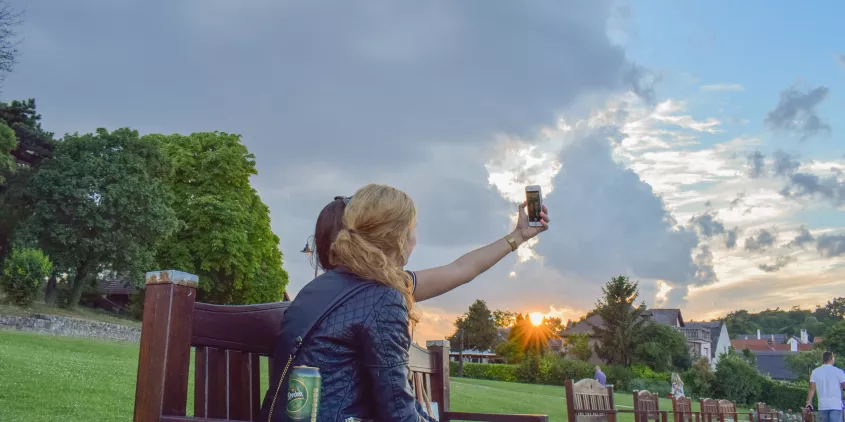 Zwei Frauen machen ein Selfie auf einer Parkbank. Im Hintergrund blauer Himmel mit Wolken und Abendröte.
