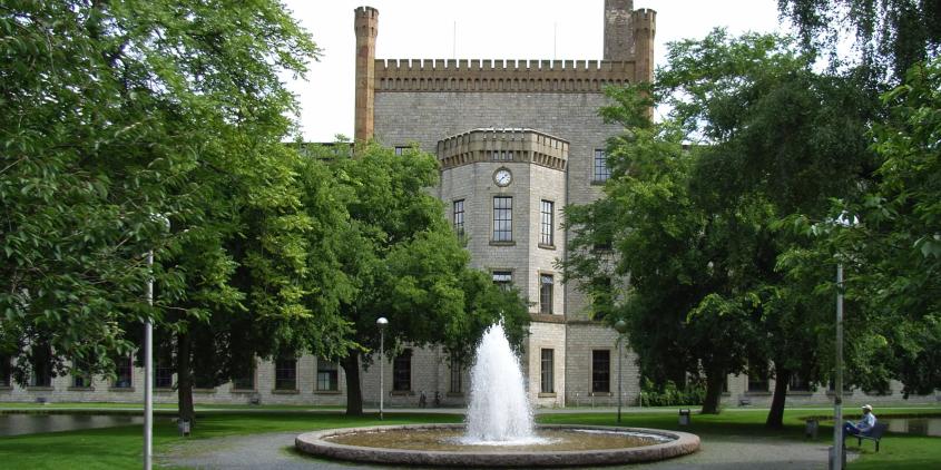 Die Ravensberger Spinnerei in der Frontalansicht. Links und rechts große Bäume. Im Fokus ein Springbrunnen direkt vor dem historischen Gebäude.