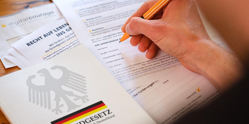 Eine Person ist dabei, die Petition zu unterzeichnen. Neben der ausgedruckten Petition liegt das Grundgesetz.