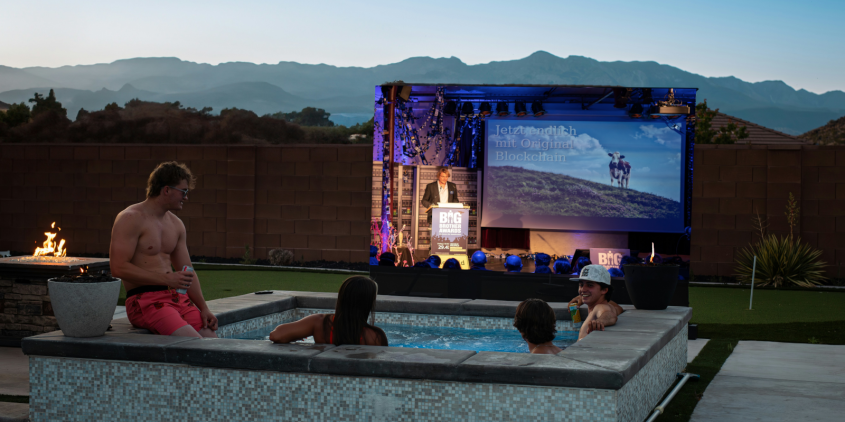 Fotomontage: Menschen in einem Whirlpool bei Dämmerung. Sie schauen auf eine Leinwand. Abgebildet ist ein Ausschnitt der BigBrotherAwards-Gala.