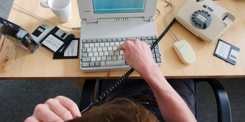 Draufsicht wie eine Person auf einem sehr alten Laptop tippt. Daneben Disketten und ein Wählscheibentelefon.