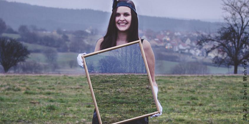 Eine Frau steht auf einem Feld und hält einen großen Spiegel vor ihrem Torso in ihren Händen. Es spiegelt nicht sich die Kamera, sondern es erscheint das Feld hinter ihr.