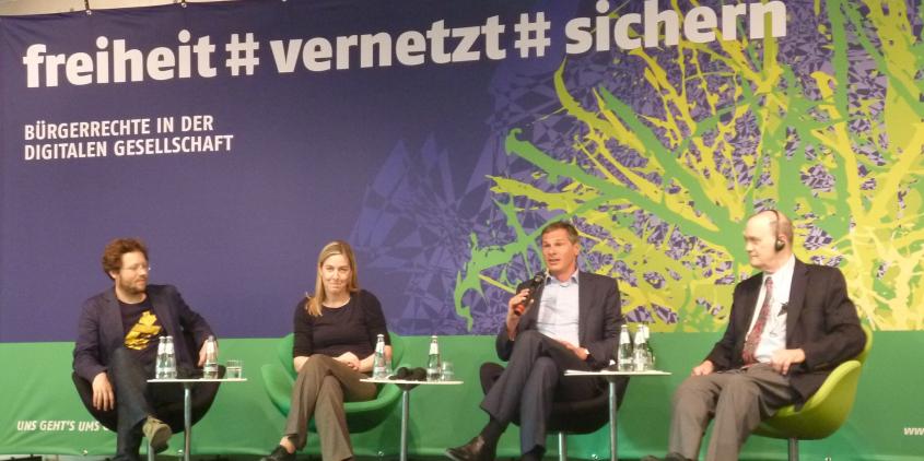 Mehrere Personen sitzen auf einem Podium. Im Hintergrund ein riesiger Banner: „freiheit #vernetzt# sichern“.