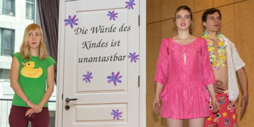 Drei Personen, zwei davon, die wie Barbies verkleidet sind neben eine Tür, auf der steht „Die Würde des Kindes ist unantastbar“.