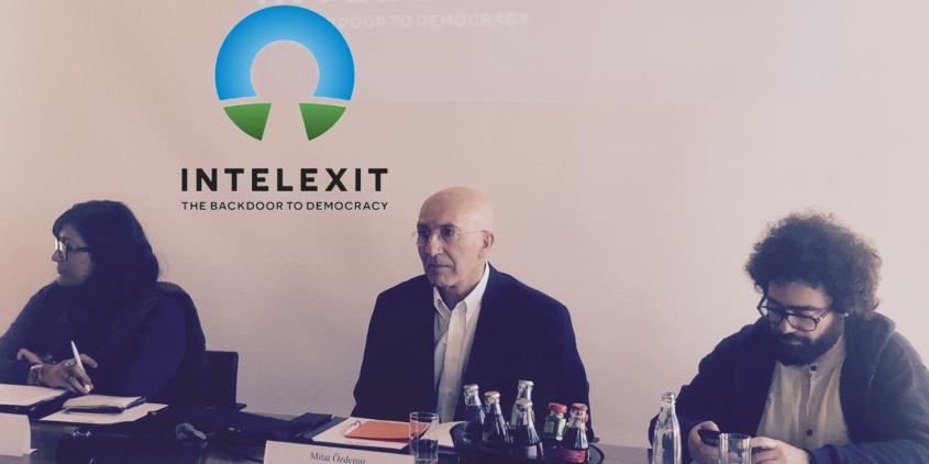 Drei Personen sitzen an einem Tisch während einer Pressekonferenz. Im Hintergrund das Intelexit-Logo.