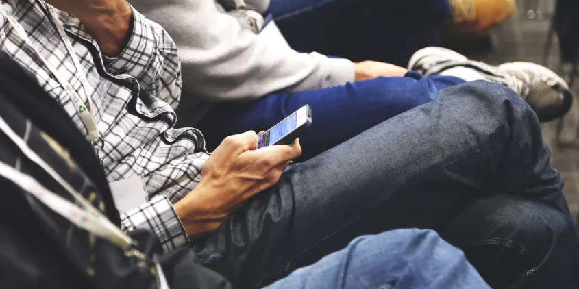 Mehrere Person sitzen in einer Reihe. Fokus auf ein Smartphone in der Hand.