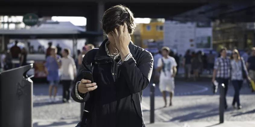 Eine Person in belebter Umgebung, der auf sein Handy schaut und sich an den Kopf fasst.