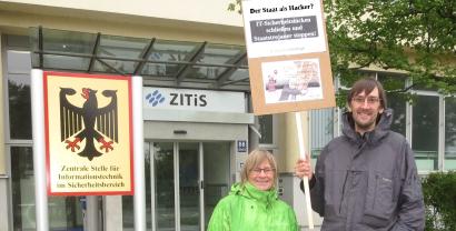 Zwei Personen mit einem Demoschild vor der Zentralen Stelle für Informationstechnik im Sicherheitsbereich. 