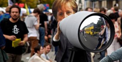 Ein riesiges Fernglas auf einer Demo aufgebaut. Dahinter steht eine Person mit einer Angela-Merkel-Maske.
