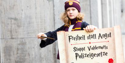 Ein Kind, das verkleidet ist wie eine Figur aus Harry Potter. In der Hand ein Schild und ein Zauberstab. Auf dem Schild steht: „Freiheit statt Angst. Stoppt Voldemorts Polizeigesetze“.