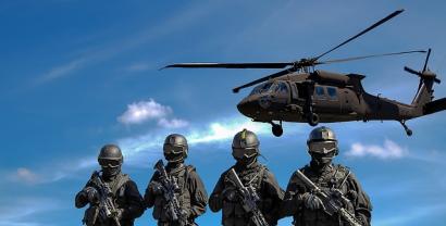 Vier Soldaten mit Gewehren stehen vor einem blauen Himmel. Im Hintergrund fliegt ein Hubschrauber.