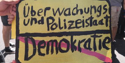 Ein Demoschild, das aussieht wie ein Ortsausgangschild (schwarze Schrift auf gelben Grund). Oben: Überwachungs- und Polizeitstaat. Unten durchgestrichen in rot: Demokratie.