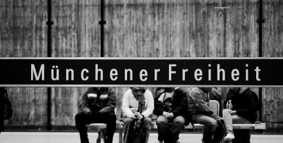 Bahnstation „Münchener Freiheit“. Das Stationsschild verdeckt die Köpfe der im Hintergrund sitzenden Menschen (Aufnahme in schwarz-weiß).