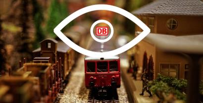 Ein großes Auge, dessen Pupille aus dem Logo der Deutschen Bahn AG besteht, überblickt einen Miniaturbahnhof.