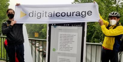 Zwei Personen halten ein Banner mit dem Logo und Schriftzug von Digitalcourage vor einem menschenhohen Smartphone-Aufsteller