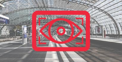 Rotes Kampagnenlogo mit einem stilisierten Auge. Im Hintergrund eine große Bahnhofshalle. 