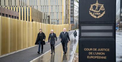 Drei Personen laufen auf die Kamera zu. Rechts im bild ist ein Schild, welches den Europäischen Gerichtshof ausweist.