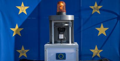 Vor einer EU-Flagge mit Sternen, die Überwachungssymbole in sich haben, steht ein Apparat. Der Apparat hat mittig eine Handyhalterung, oben eine Alarmleuchte und unten ein Gehäuse aus Plexiglas mit Metallprofilen.