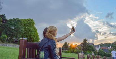 Zwei Frauen machen ein Selfie auf einer Parkbank. Im Hintergrund blauer Himmel mit Wolken und Abendröte.