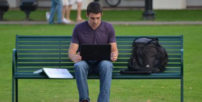Eine Person sitzt auf einer Parkbank. Auf dem Schoß steht ein Laptop, auf der Bank liegt ein aufgeschlagenes Buch.
