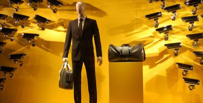 Eine Schaufensterpuppe in Anzug mit zwei Taschen vor gelben Hintergrund, auf die sehr viele Überwachungskameras gerichtet sind.