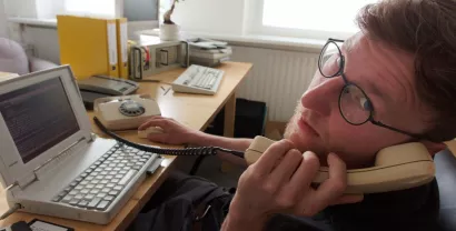 Ein Digitalcourage-Mitarbeiter vor einem alten PC mit Schnurtelefon, direkt in die Kamera schauend.