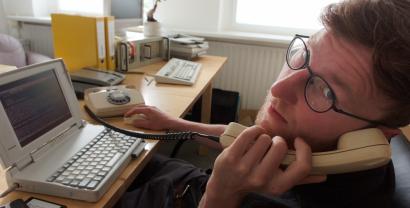 Ein Digitalcourage-Mitarbeiter vor einem alten PC mit Schnurtelefon, direkt in die Kamera schauend.