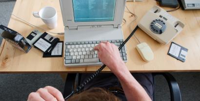 Ein Digitalcourage-Mitarbeiter tippt auf einem sehr alten Laptop. In der Hand ein Schnurtelefon.