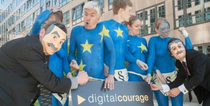 Mehrere nackt Menschen, die wie die Europa-Flagge angemalt sind (blau mit gelben Sternen). Daneben Menschen mit Masken. SIe halten ein Schild von Digitalcourage in den Händen.