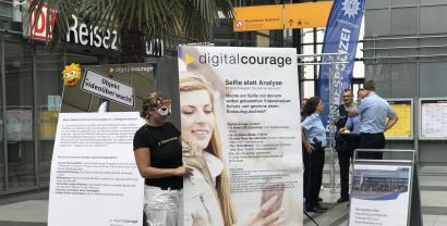 Leena Simon mit einer Maske neben einem Plakat „Selfie statt Analyse“. Im Hintergrund sind mehrere Polizist.innen. Aufnahmeort: Ein Bahnhof.