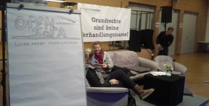 Ein Flipchart mit dem Text "Open Sofa – Lesen gegen Überwachung". Im Hintergrund zwei Personen auf einem Sofa sitzend.