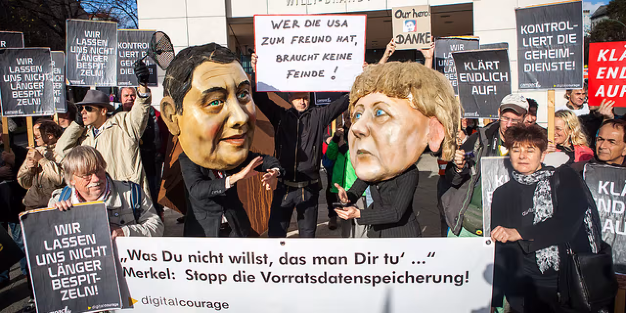 Aktivist.innen mit diversen Bannern. Im Hintergrund Pappmaché-Köpfe von Angela Merkel und Sigmar Gabriel.