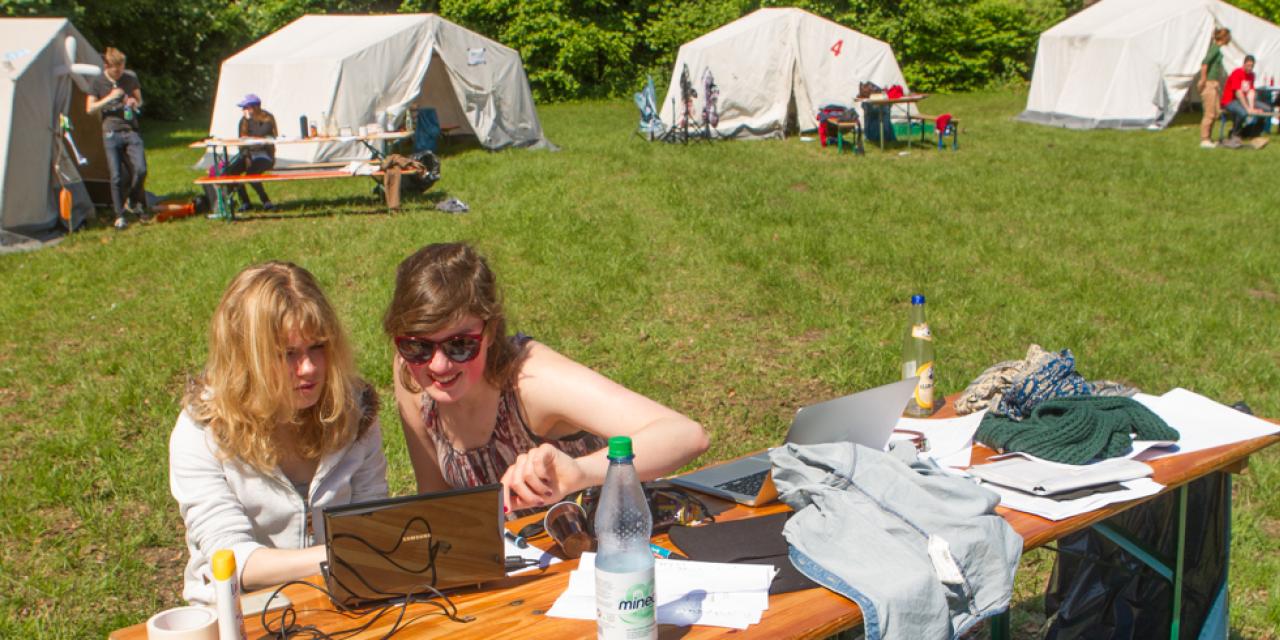 Zwei Personen sitzen an einem Tisch vor einem Laptop. Im Hintergrund stehen Zelte.