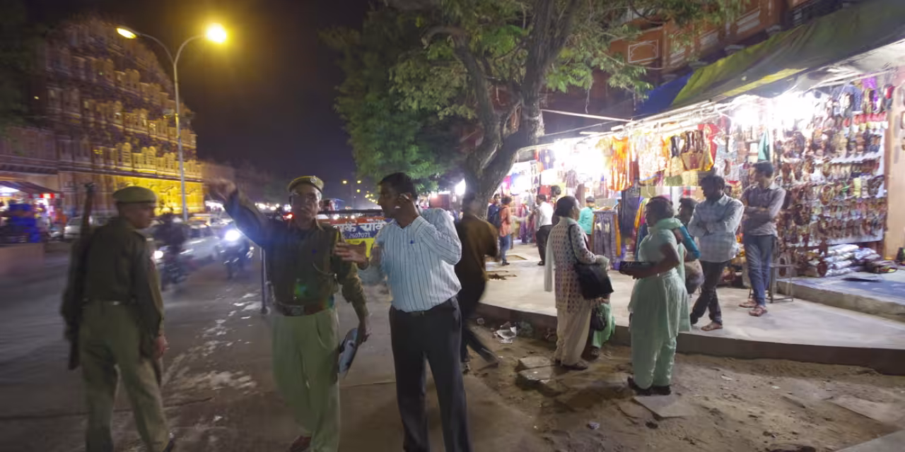 Polizei in Indien zeigt in eine Richtung. Daneben steht eine Person. Im Hintergrund sind Straßengeschäfte.