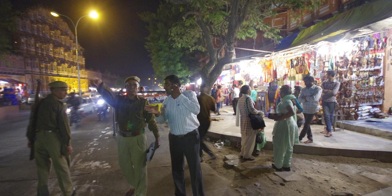 Polizei in Indien zeigt in eine Richtung. Daneben steht eine Person. Im Hintergrund sind Straßengeschäfte.