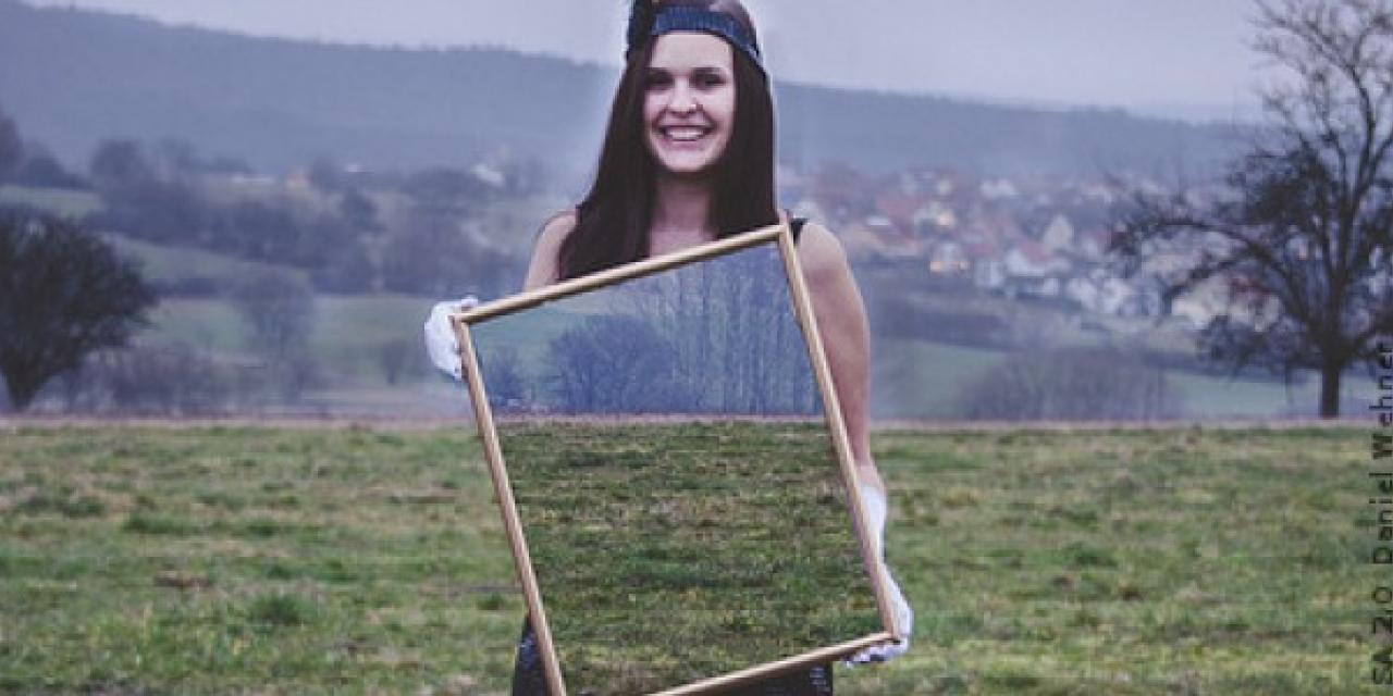 Eine Frau steht auf einem Feld und hält einen großen Spiegel vor ihrem Torso in ihren Händen. Es spiegelt nicht sich die Kamera, sondern es erscheint das Feld hinter ihr.
