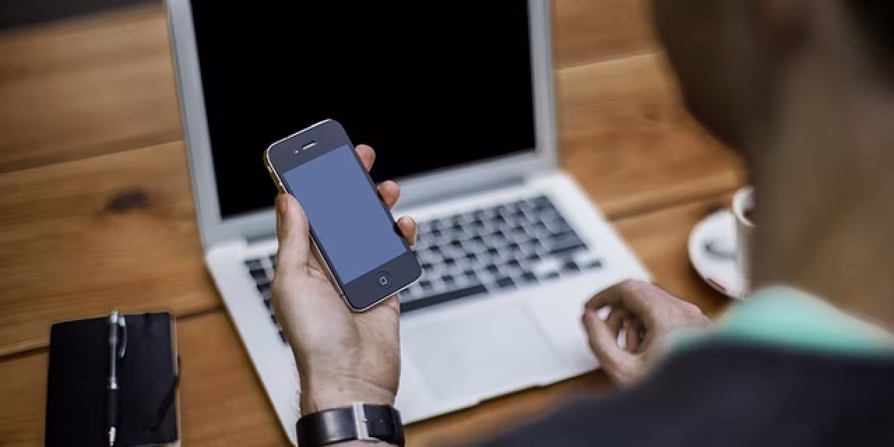 Eine Person an einem Laptop, die gleichzeitig auf ein Smartphone schaut.