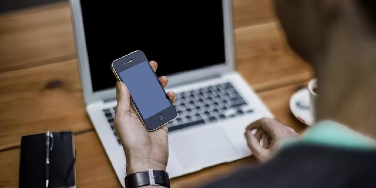 Eine Person an einem Laptop, die gleichzeitig auf ein Smartphone schaut.