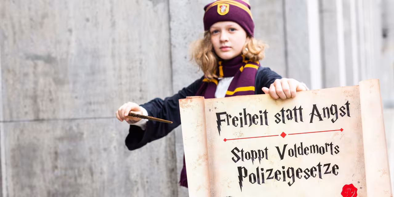 Ein Kind, das verkleidet ist wie eine Figur aus Harry Potter. In der Hand ein Schild und ein Zauberstab. Auf dem Schild steht: „Freiheit statt Angst. Stoppt Voldemorts Polizeigesetze“.