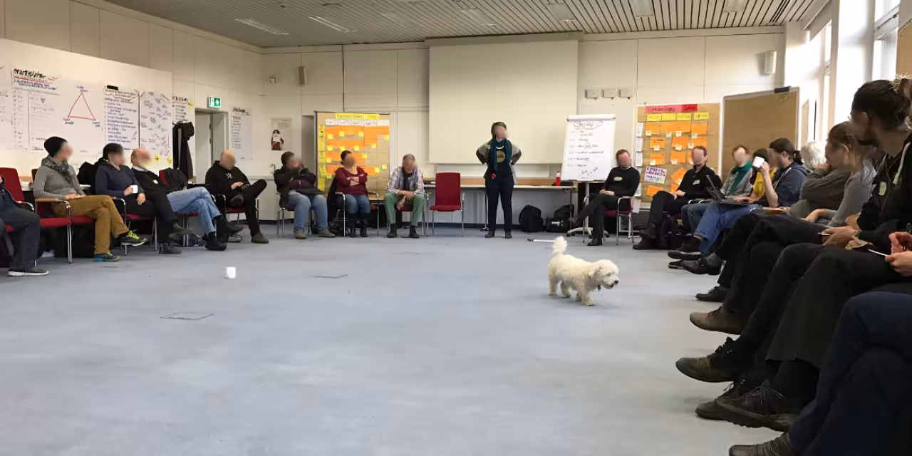 Ein Sitzkreis mit vielen Menschen, eine Person steht an einer Flipchart. 