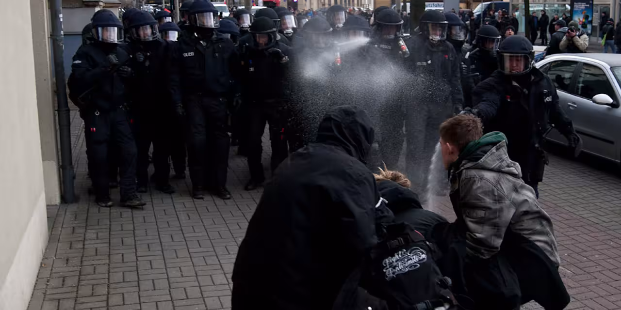 Drei Demonstrant.innen werden von mehreren Polizist.innen mit Pfefferspray besprüht.
