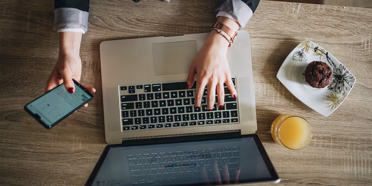 Draufsicht eines Laptops mit einer Person, die tippt und gleichzeitig ein Smartphone in der Hand hält.