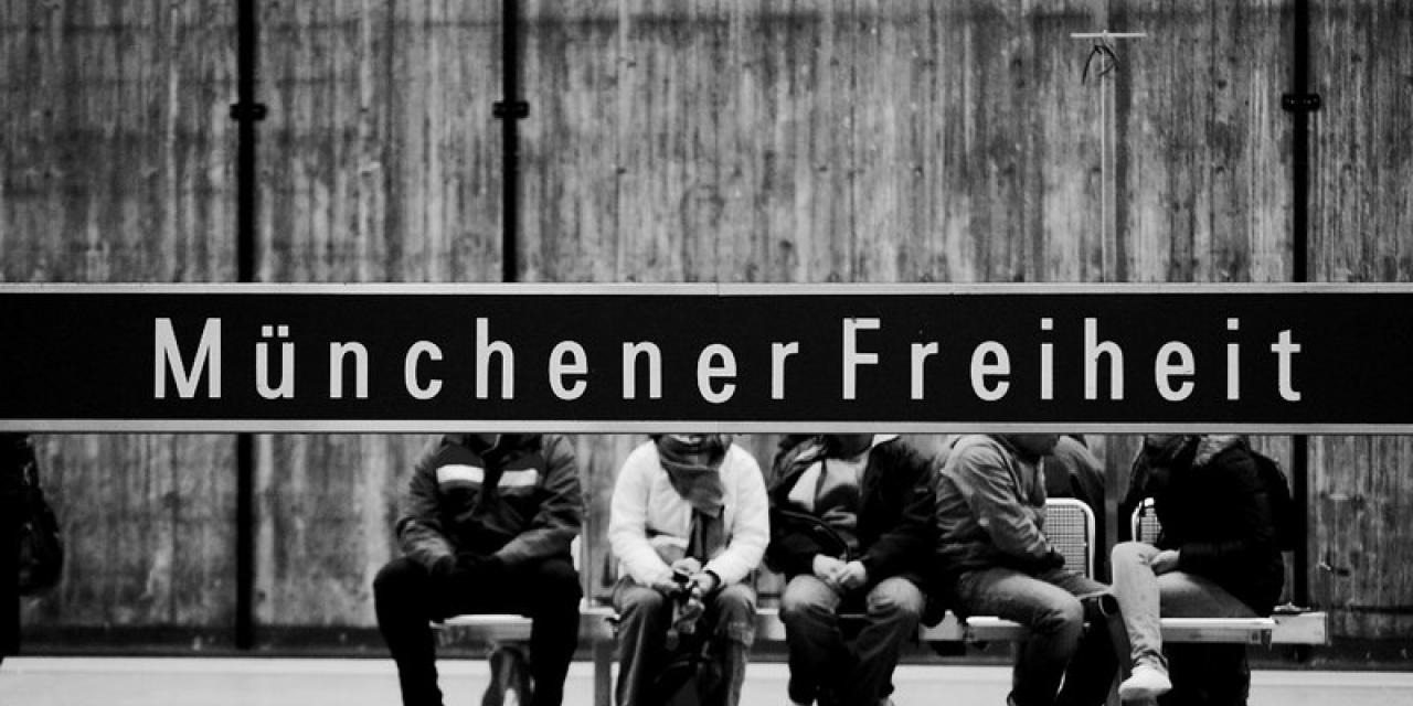 Bahnstation „Münchener Freiheit“. Das Stationsschild verdeckt die Köpfe der im Hintergrund sitzenden Menschen (Aufnahme in schwarz-weiß).