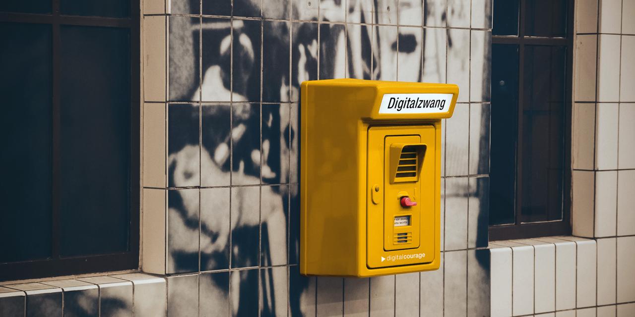 Montage: Ein gelber Kasten an einer Hauswand. Darauf steht „Digitalzwang“.