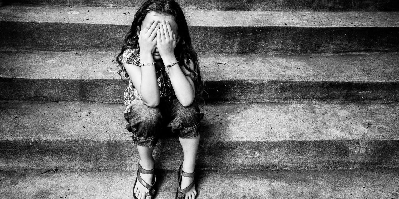 Ein Kind sitzt auf einer Treppe und hält sich die Hände vor das Gesicht (schwarz/weiß).