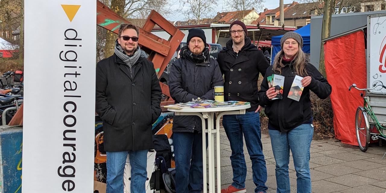 Vier Mitglieder der Ortsgruppe Bremen stehen lächelnd um einen Stehtisch neben einem Digitalcourage-Banner auf einem Markt und zeigen stolz Flyer her