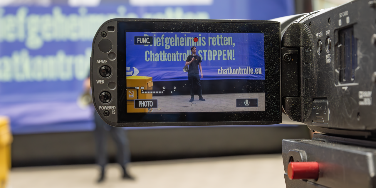 Ein Mensch ist auf dem Bildschirm einer Videokamera zu sehen. Er steht, mit einem Megaphon bewaffnet, vor einem Protestbanner.